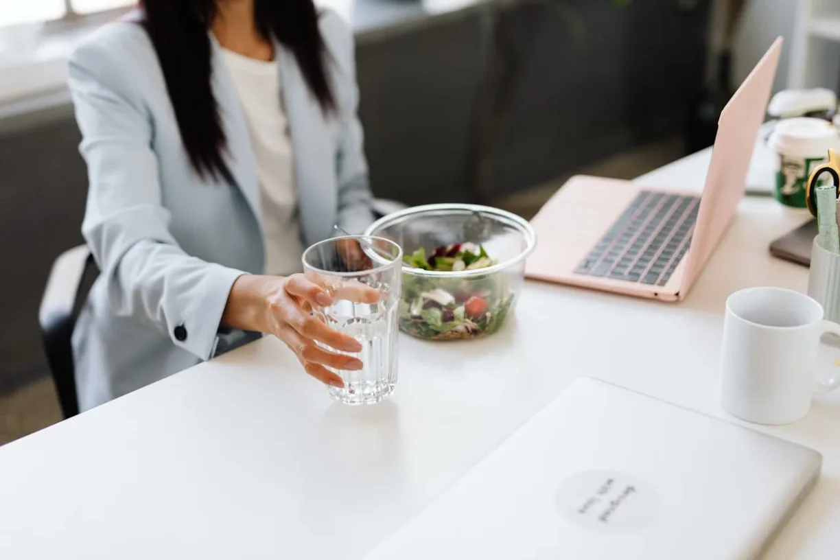 Office worker reaching for a glass of water beside a laptop and healthy lunch at a desk.