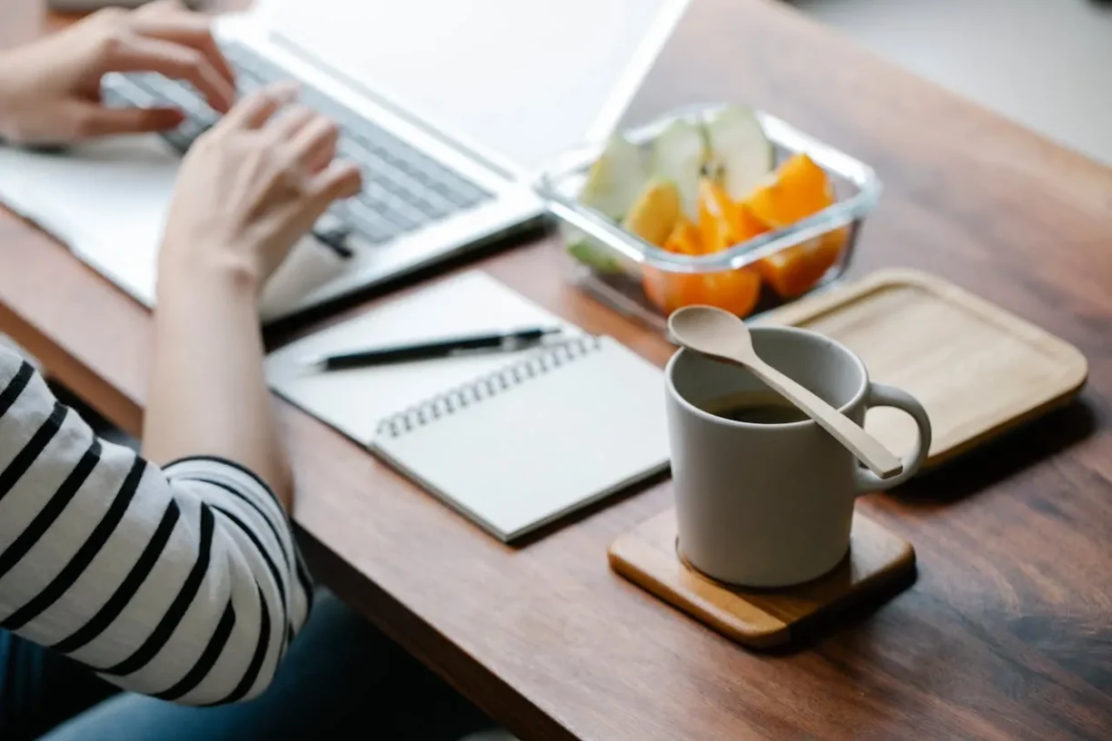 Person working on a laptop with coffee and fruit, representing a balanced and productive work routine.